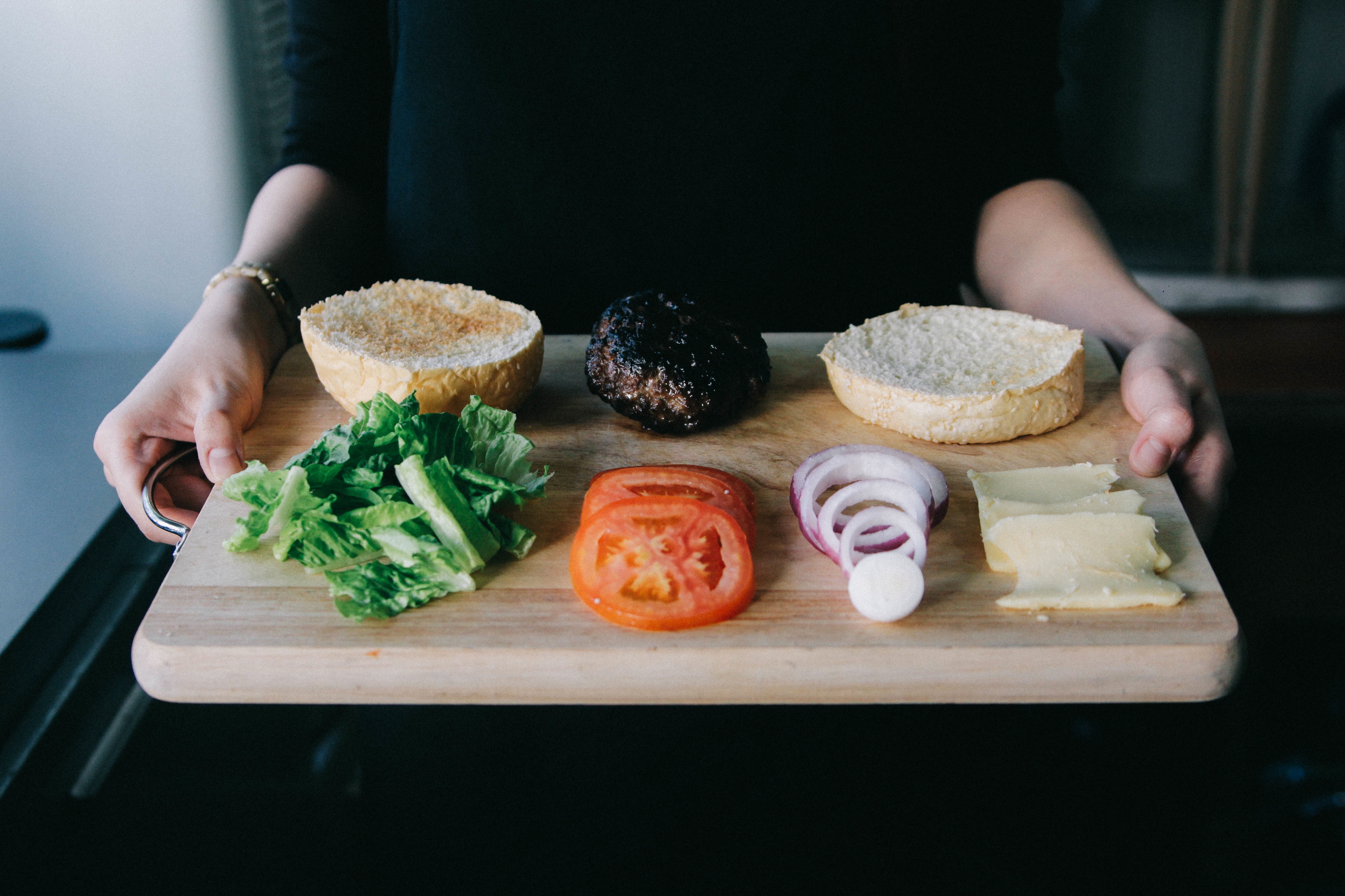 Woman holding a cutting board with ground beef, a hamburger bun, lettuce, tomato, and onion.